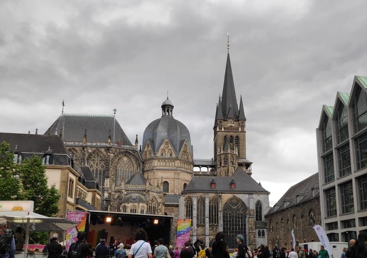 Im Hintergrund, die Rückseite des Aachener Doms. Im Vordergrund der CSD Aachen 2025 mit Bühne. Bewölkter Himmer in Abendstimmung.